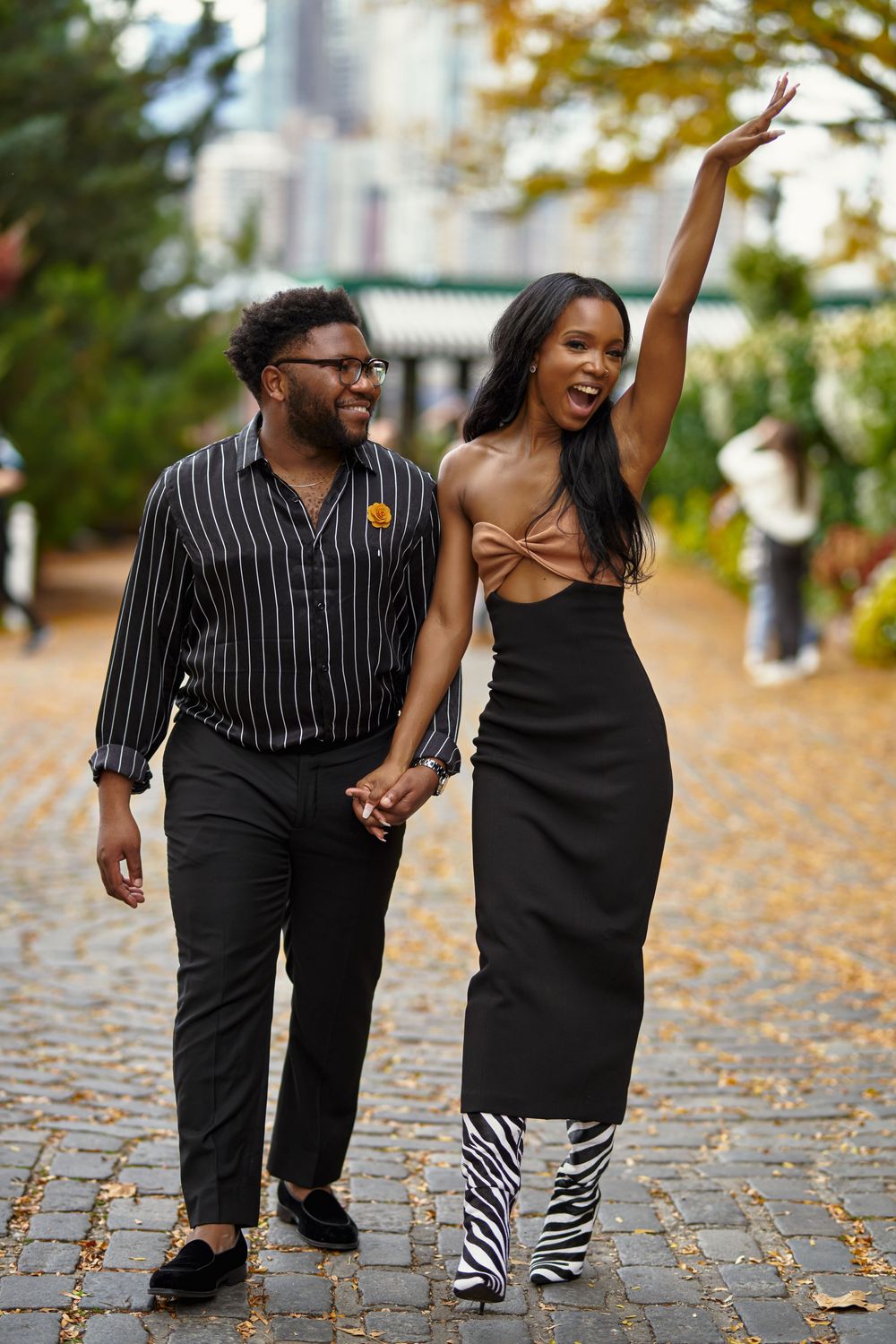 A couple walking together on a cobblestone path in autumn, wearing stylish black outfits with zebra print boots at Brooklyn Bridge