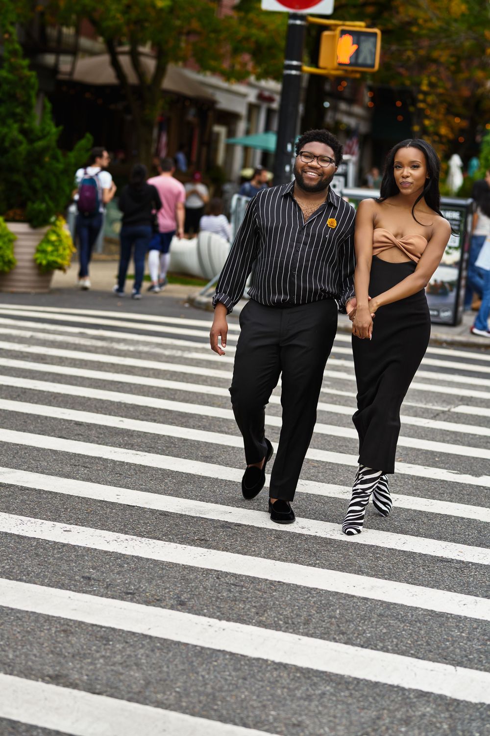 A stylishly dressed couple walks across a crosswalk in an urban setting with a traffic light visible. at Brooklyn Bridge