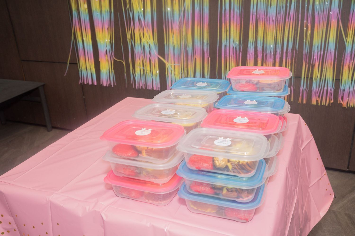 Colorful rainbow striped curtains hanging over rectangular plastic containers on a pink tablecloth at an indoor event.