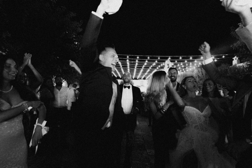 Black and white photo of people dancing and celebrating at a nighttime wedding reception under string lights.