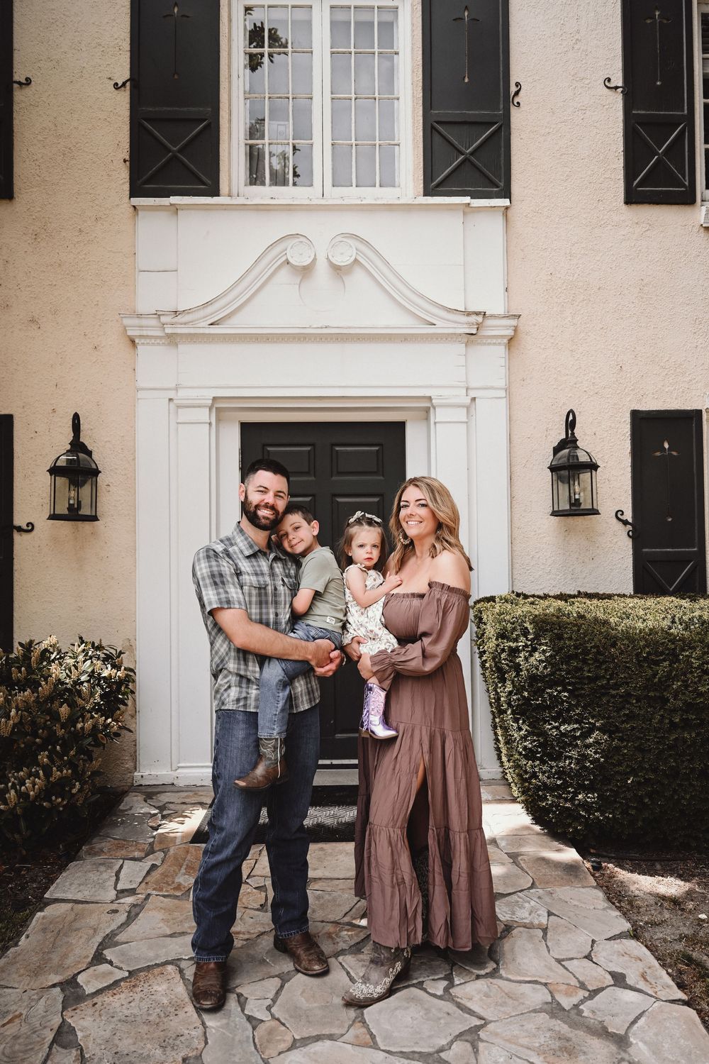 A happy family of four poses in front of an elegant home with black shutters and a stone pathway.