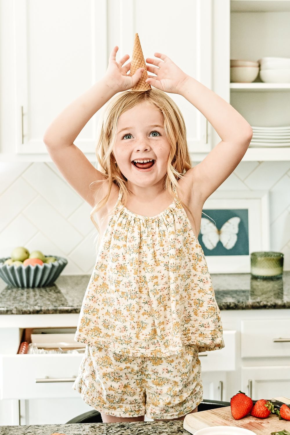 Cheerful young blonde girl holding an ice cream cone on her head, laughing in a bright white kitchen.