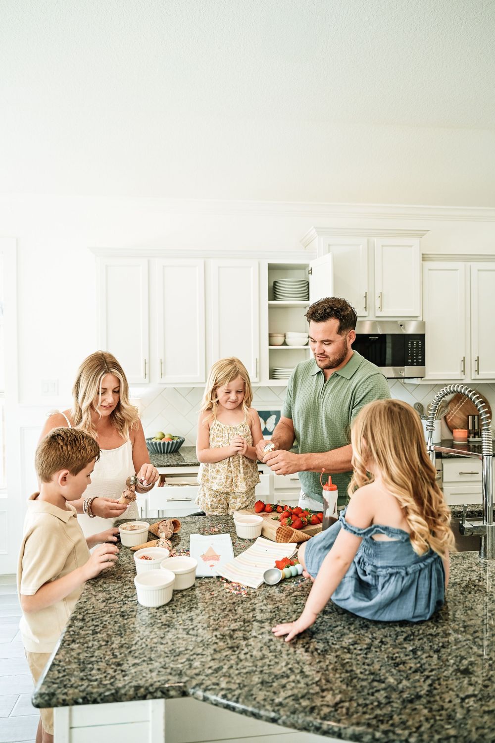 Family of five preparing food together at a granite kitchen island in a bright white kitchen.