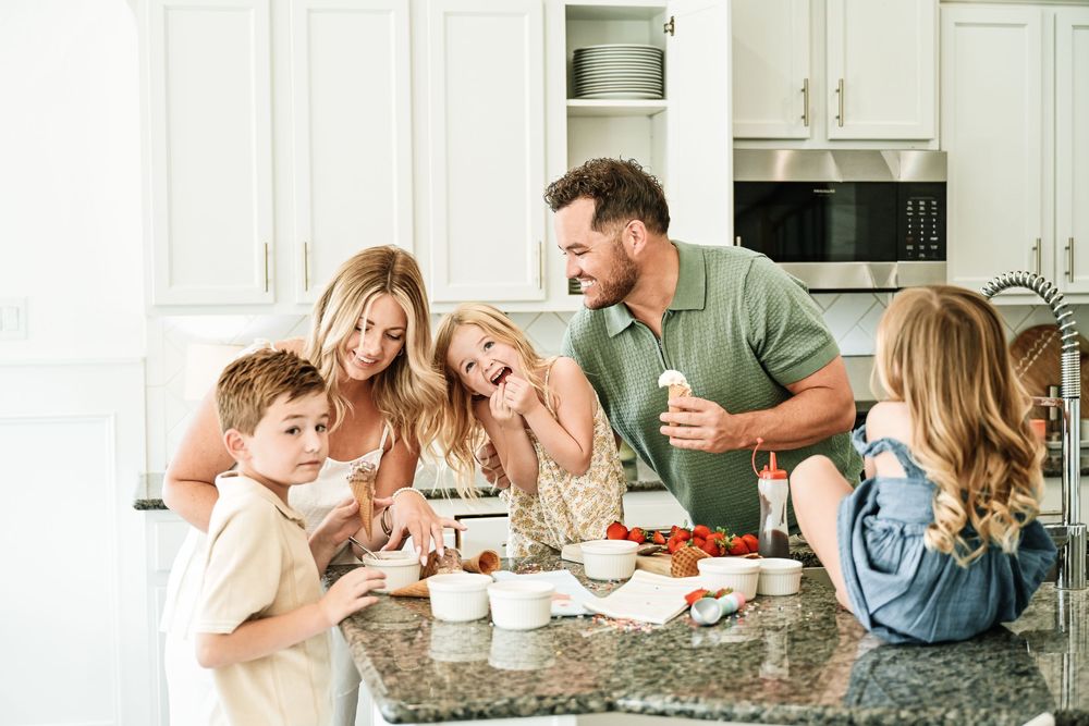 Family of five enjoying a fun cooking moment together in a bright white kitchen with fresh strawberries on the counter.