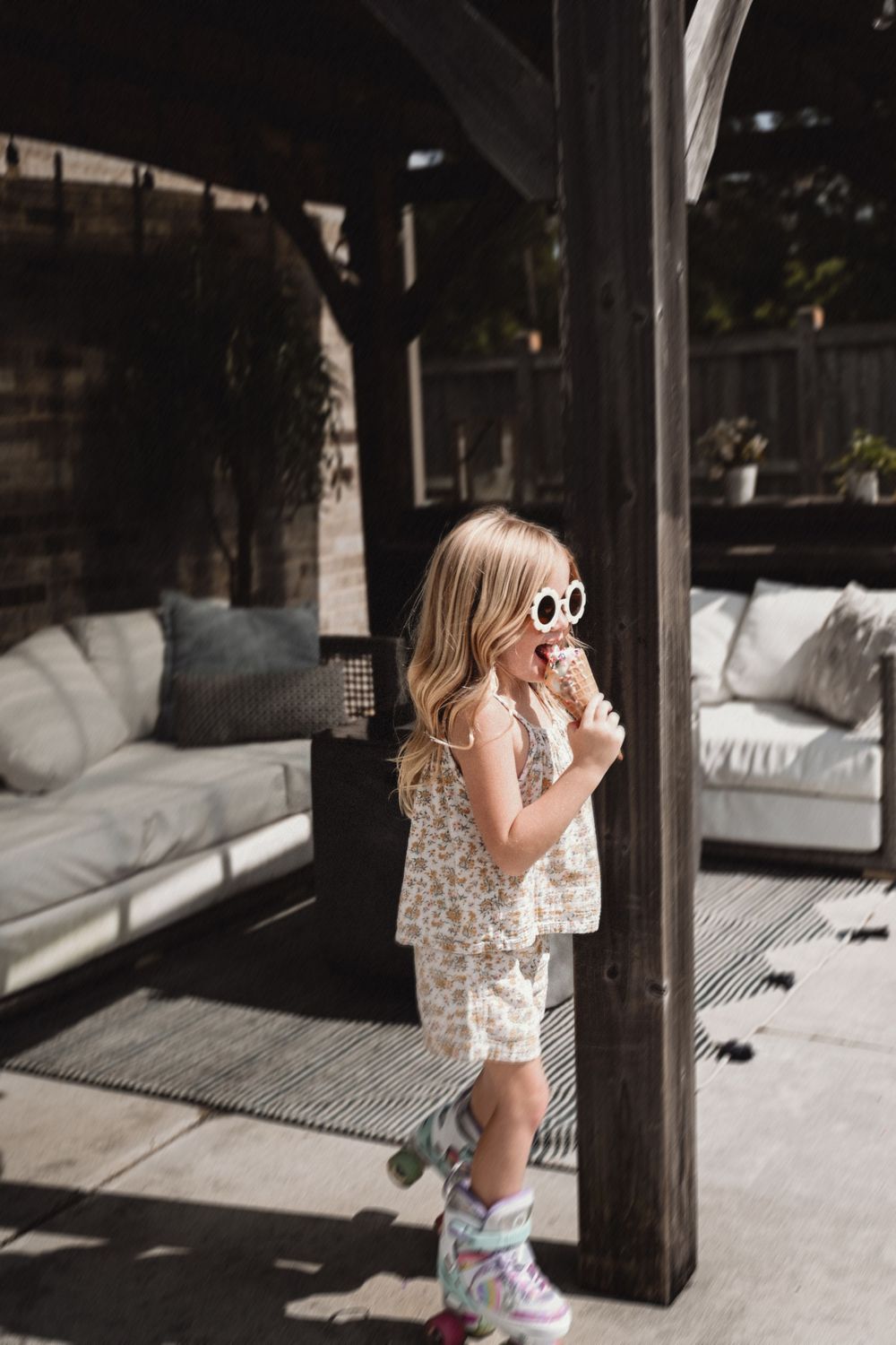Young girl in floral outfit and white sunglasses eating ice cream cone on outdoor patio.