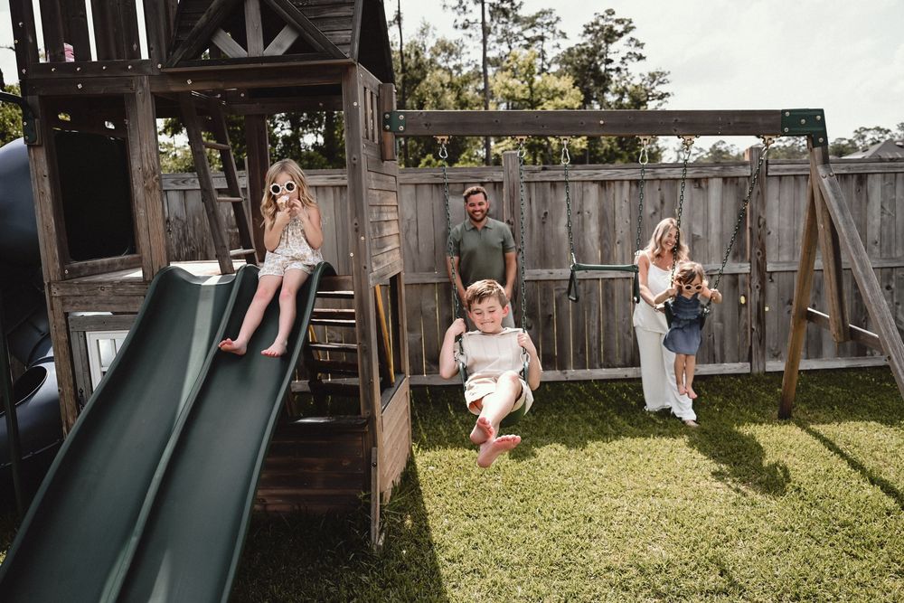 Family enjoying backyard playset with children on slide and swing while parents watch on sunny day.