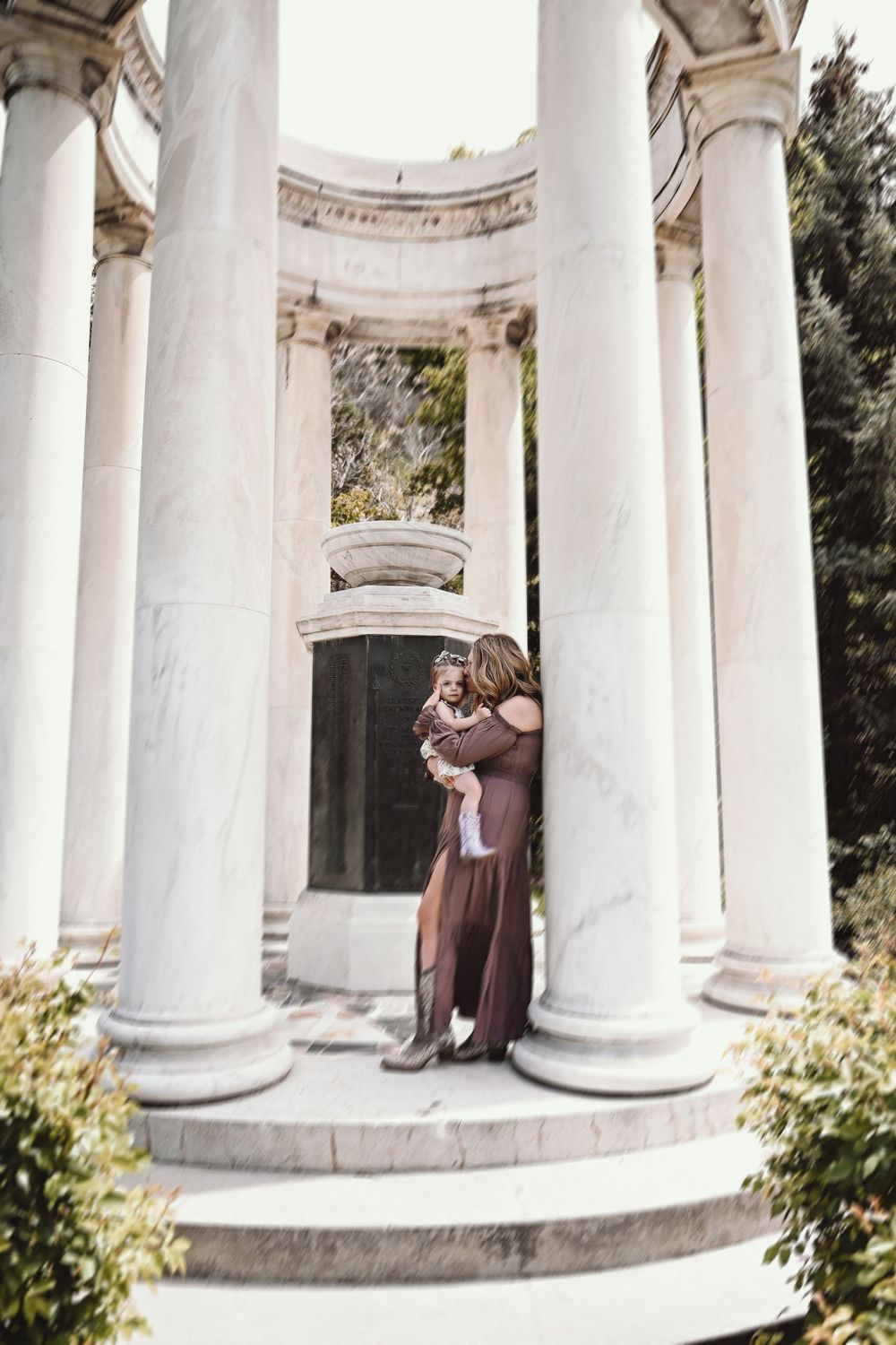 Woman in brown dress holds baby amid white marble columns and steps of a classical rotunda structure outdoors.