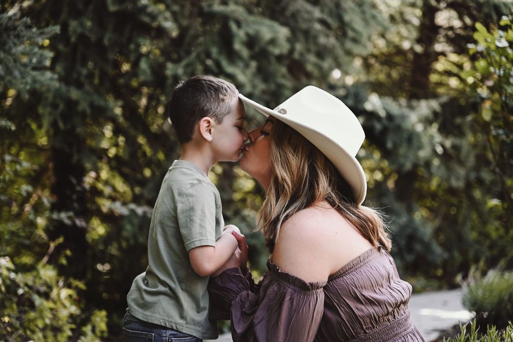 A mother in a wide-brim hat shares a tender kiss with her young son outdoors among lush green trees.