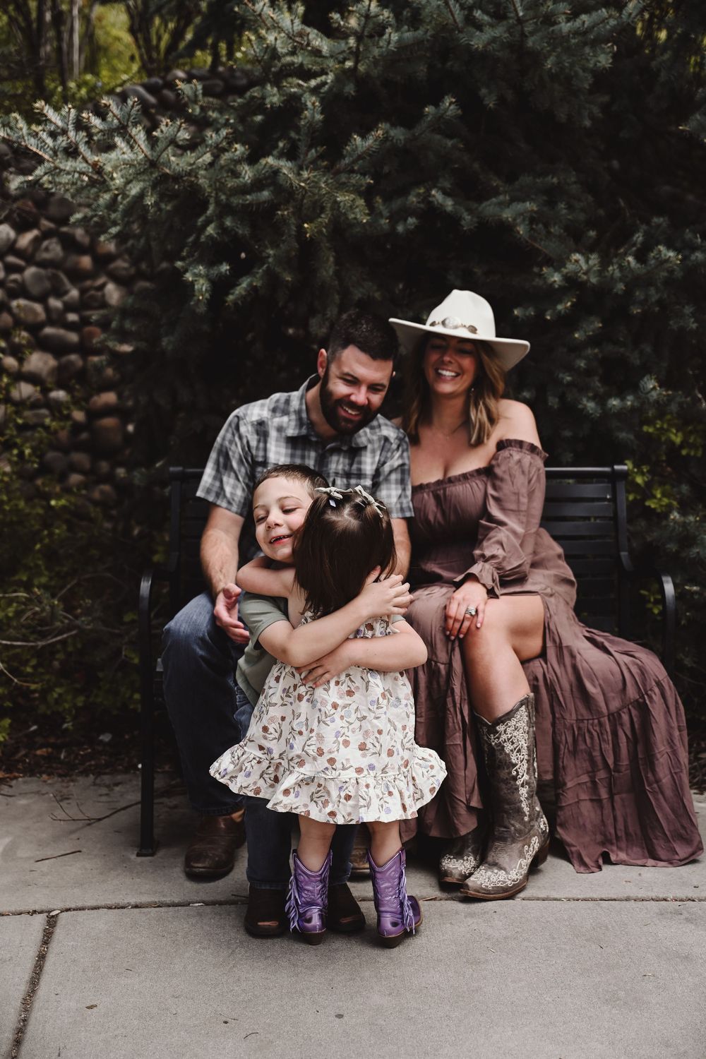 Family of four sitting on a bench outdoors, parents smiling as two young girls hug, surrounded by lush greenery.