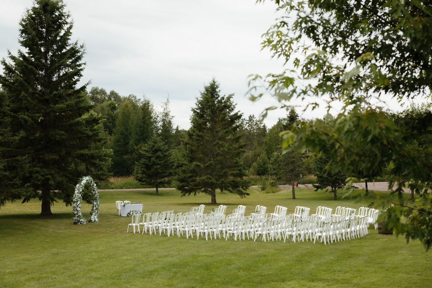 Empty white chairs arranged in rows on a green lawn for an outdoor wedding ceremony with trees in the background.