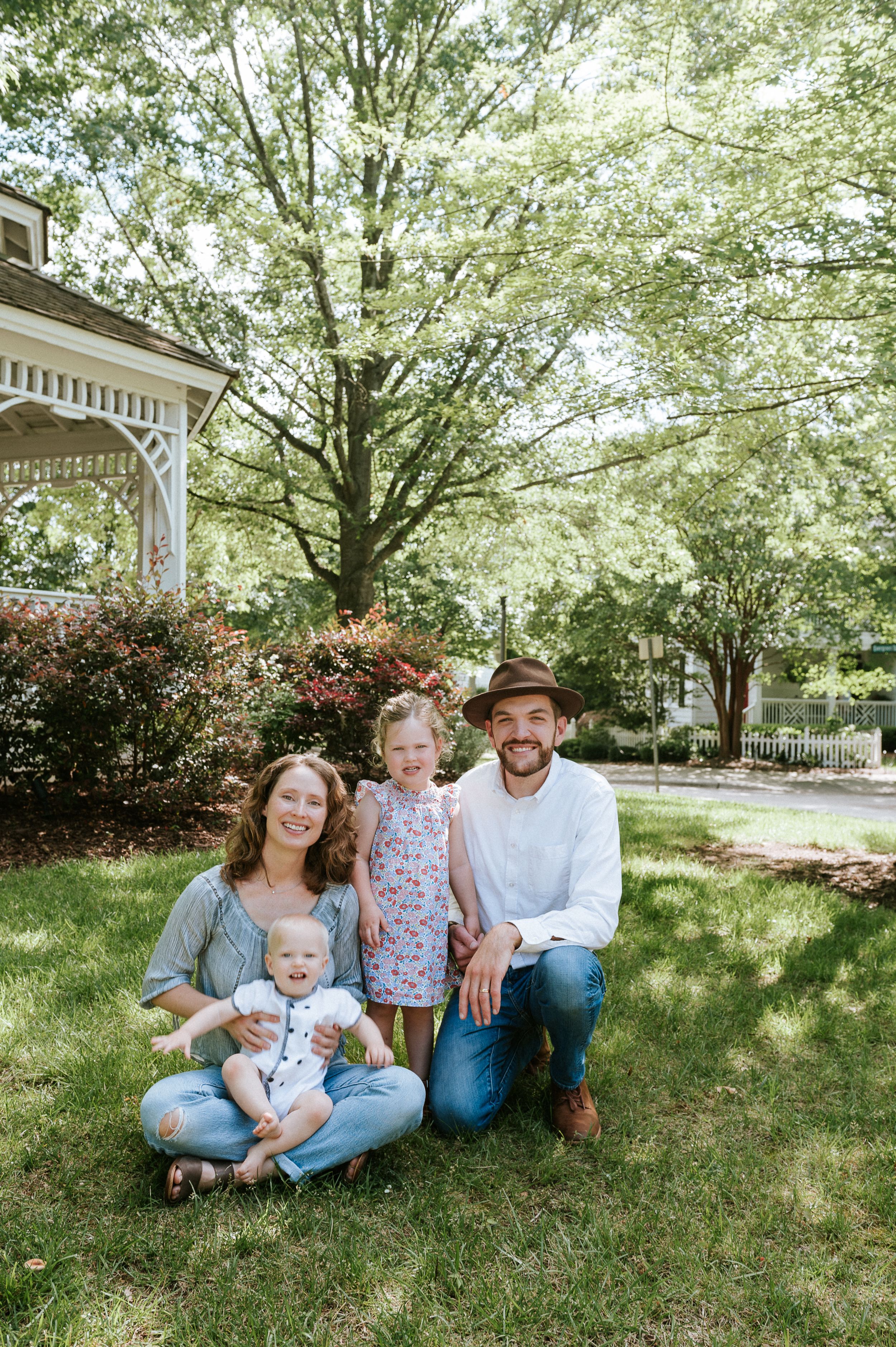 Family lounging on grass under shade trees beside white farmhouse with blooming shrubs.