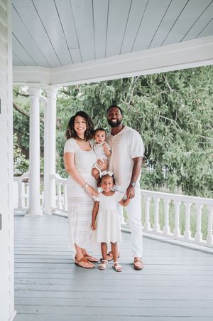 Family wearing matching white outfits poses together on rustic white porch with columns.