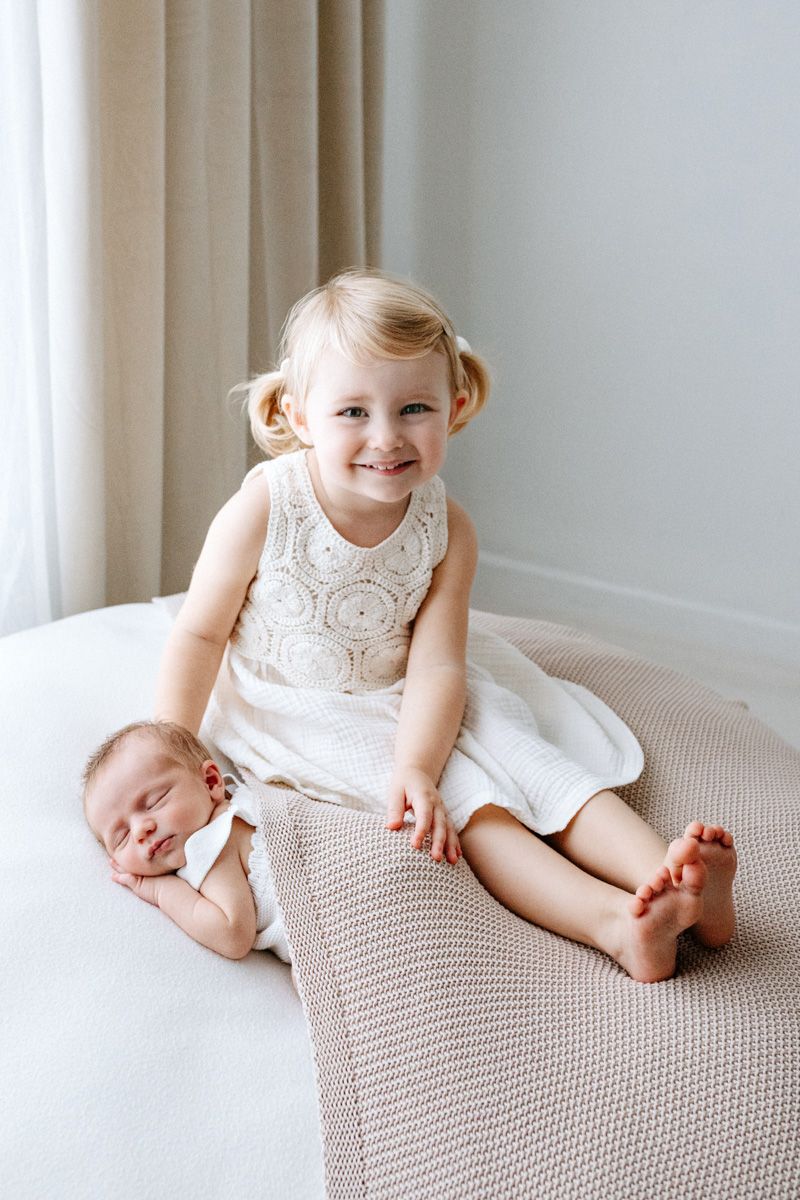 A small child in a white lace dress sits and smiles on a textured beige blanket sat next to her newborn baby brother laying on his front.