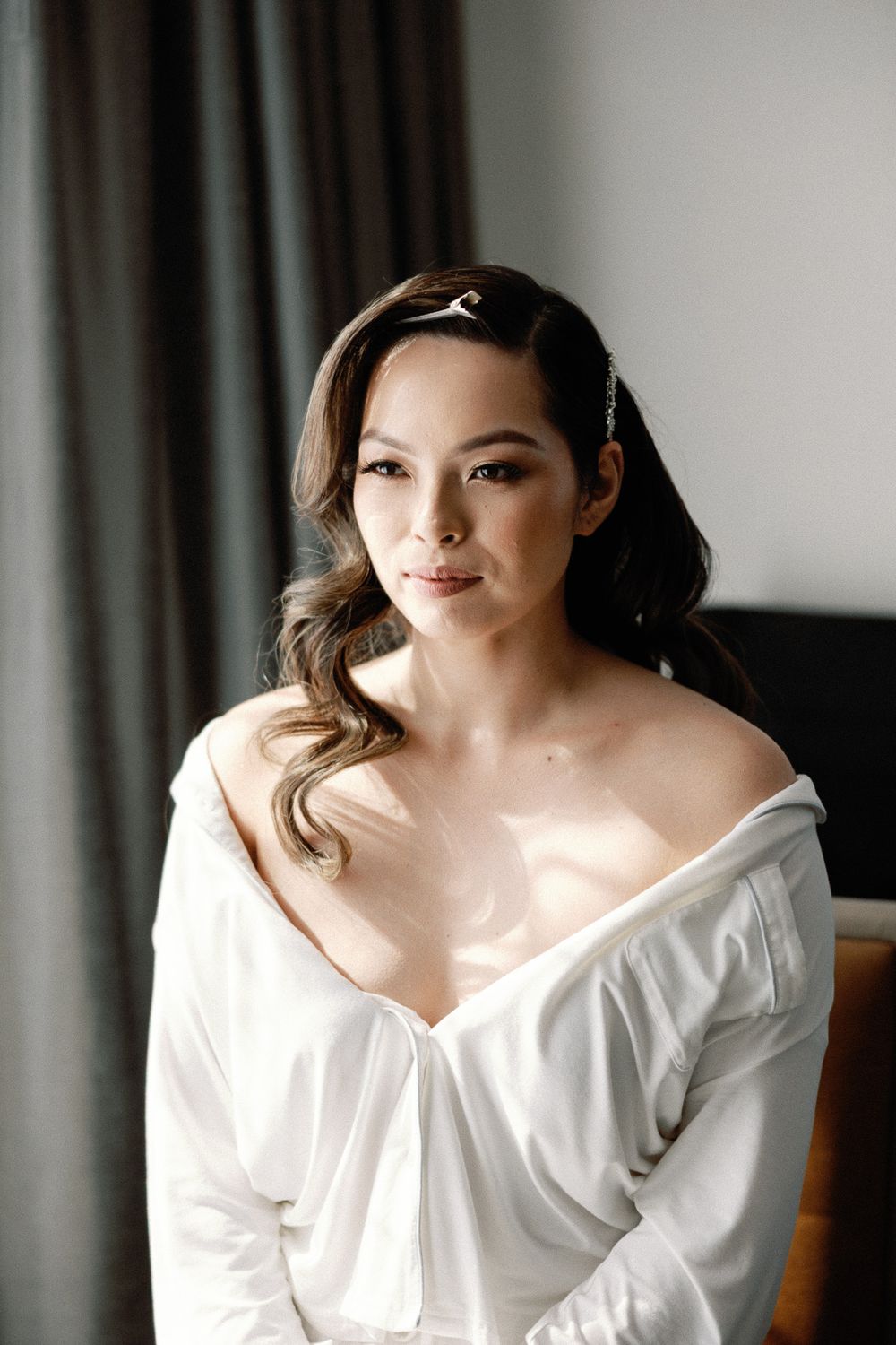 Beautiful brunette bride in white off-shoulder robe with hair accessory poses by window before wedding.