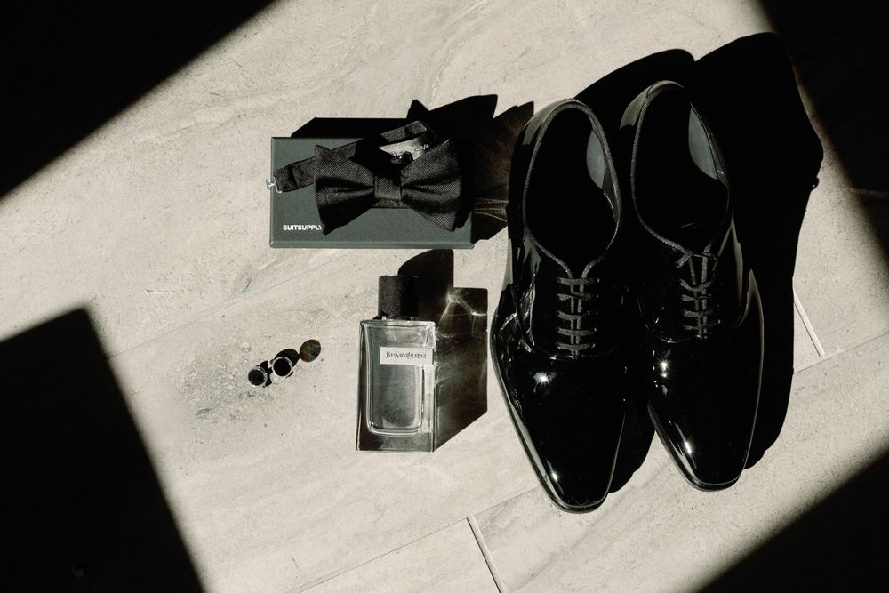Black patent leather oxford shoes, bow tie, cufflinks, and cologne arranged on a marble floor for a groom.