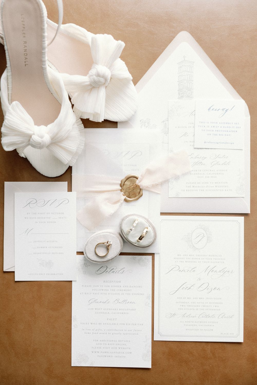 Elegant white wedding stationery suite with rings, wax seal, ribbon, and bridal heels on a neutral background.