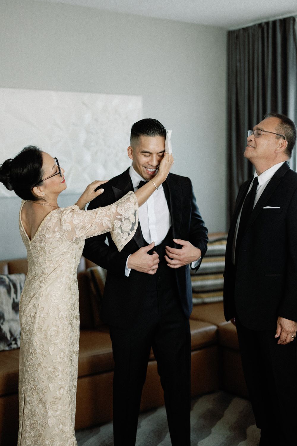 A groom in a black tuxedo smiles as his mother touches his face and his father stands proudly beside him.