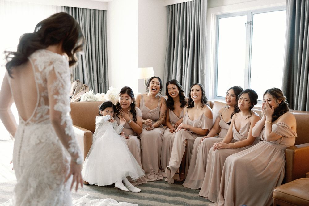 Bridesmaids in blush dresses react with joy as the bride in a lace gown reveals her look before the wedding.