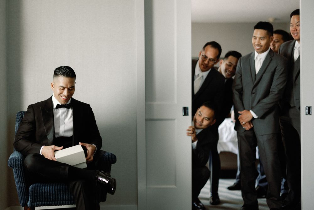 A groom reads vows in a chair while groomsmen in tuxedos peek curiously through a cracked door behind him.