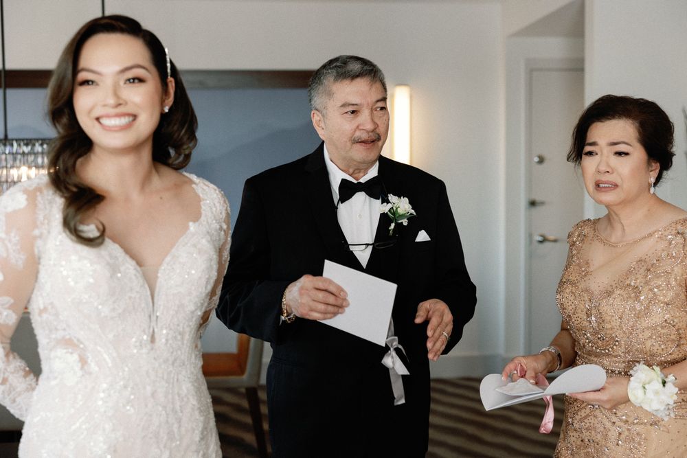 Bride in lace gown smiling with her parents holding papers at a wedding venue.