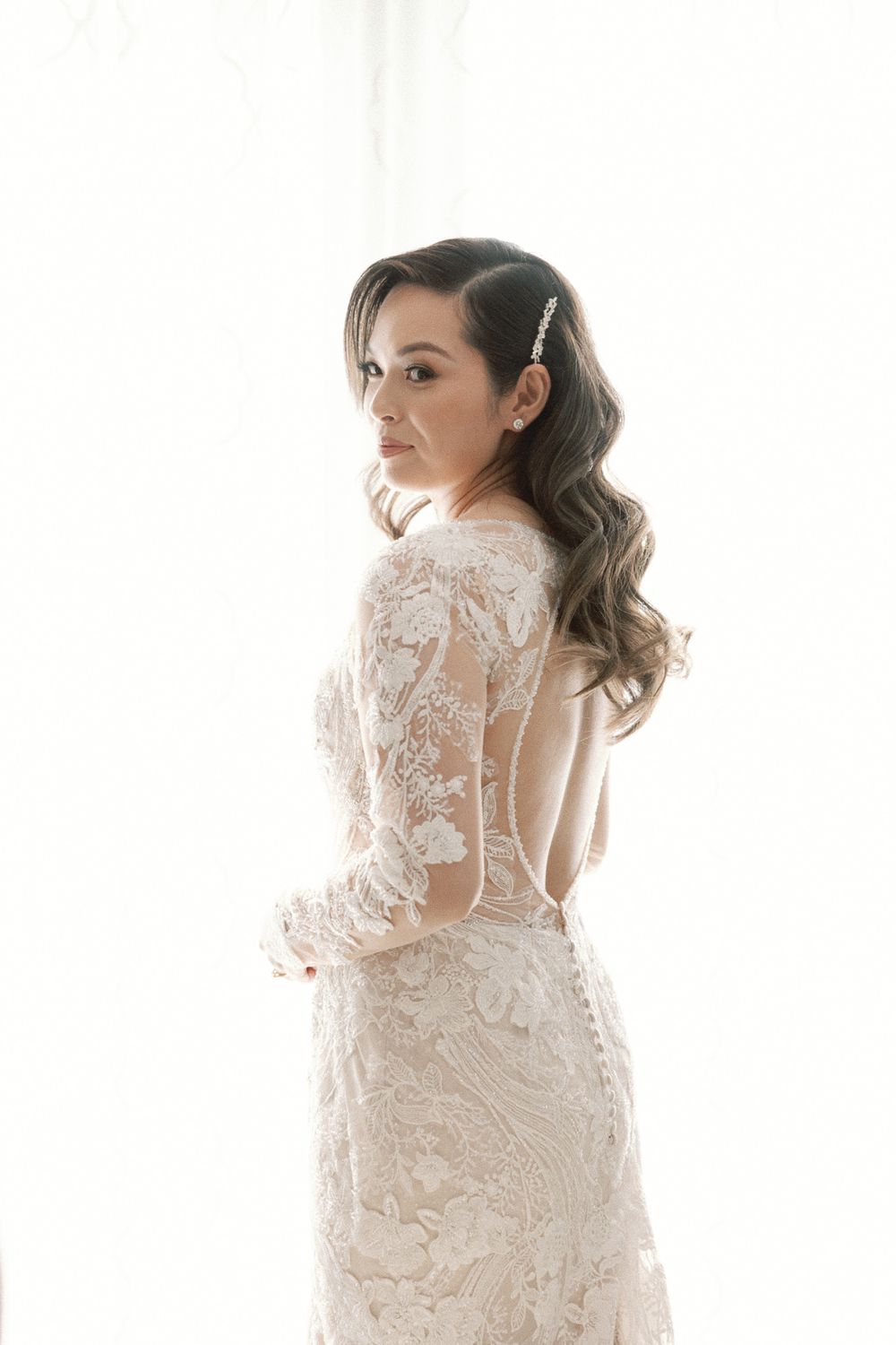 Elegant bride in backless lace long-sleeve wedding gown with crystal hair accessory poses by bright window.