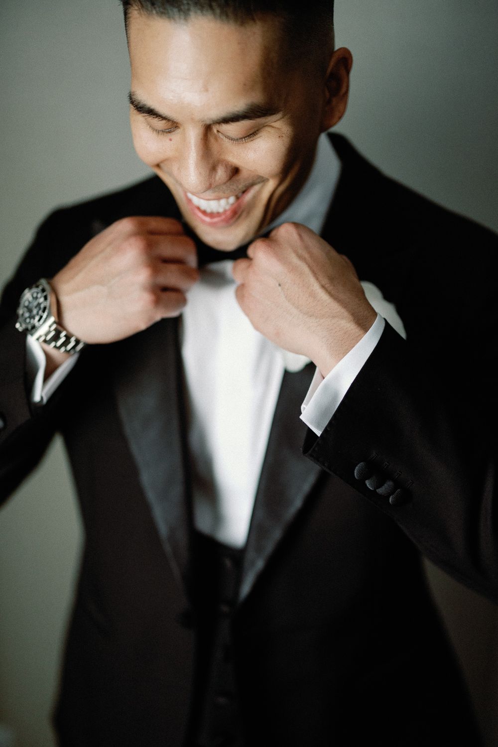 A smiling groom in a black tuxedo adjusts his bow tie, wearing a silver watch on his wrist.