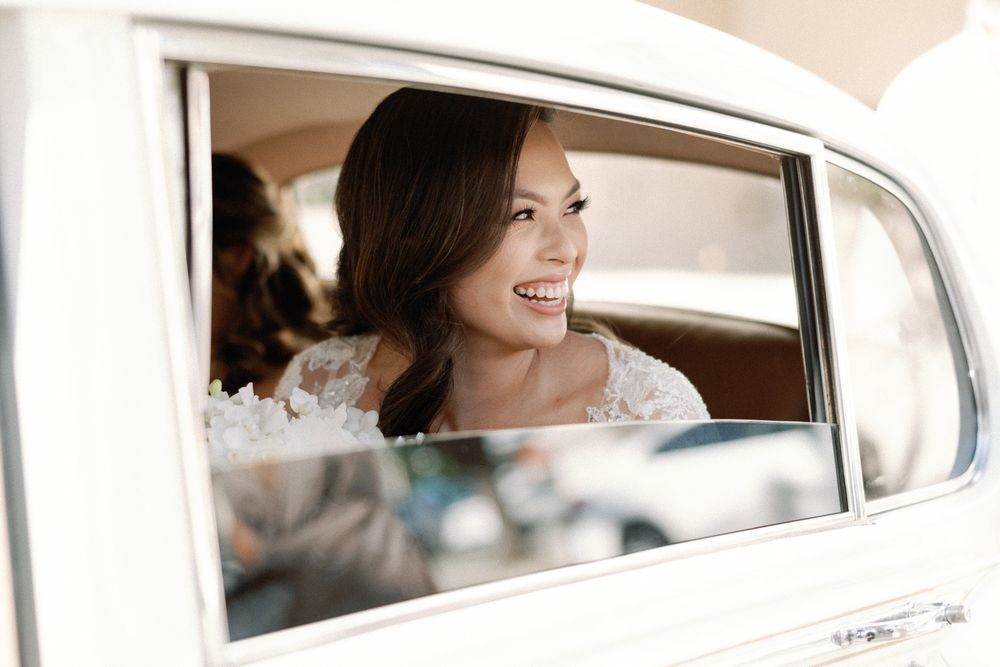 Smiling bride in lace wedding dress sitting in a vintage white car on her wedding day.