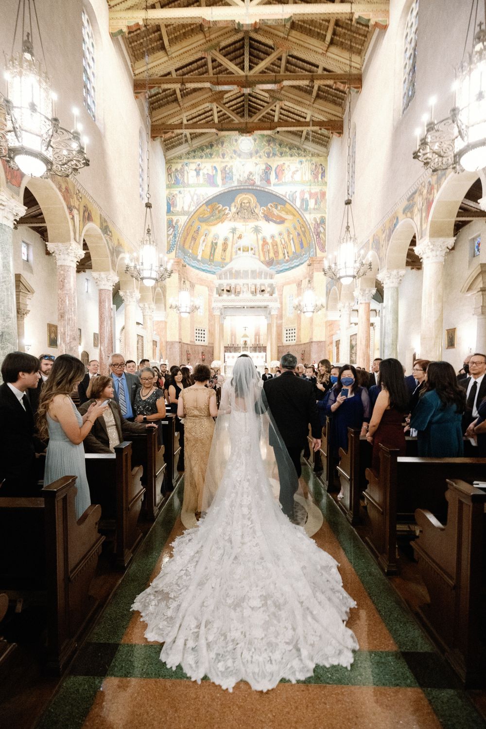 Bride in cathedral-length veil walking down ornate church aisle with wedding guests.