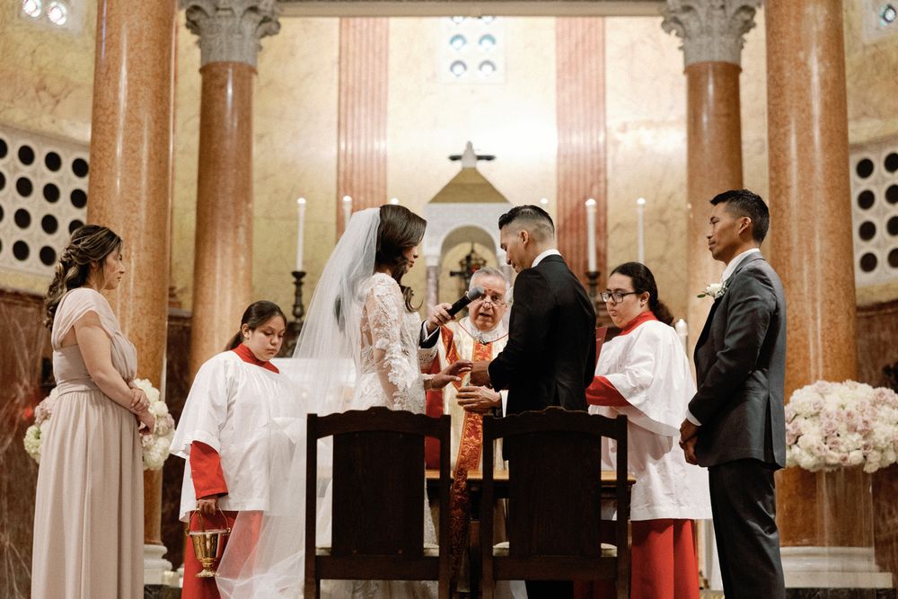 Couple exchanges vows at a Catholic church altar surrounded by wedding party and officiants.