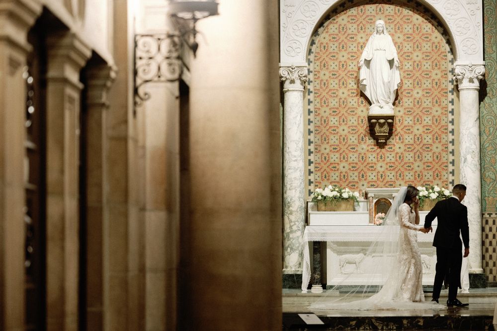 Bride and groom standing at church altar beneath a statue of the Virgin Mary.