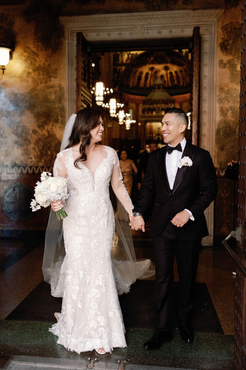 Newlyweds holding hands and exiting a grand church with ornate interior decor.