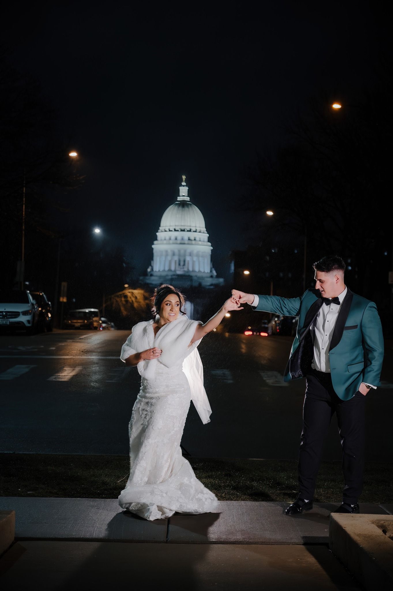 A couple poses dramatically in front of an illuminated state capitol building at night.
