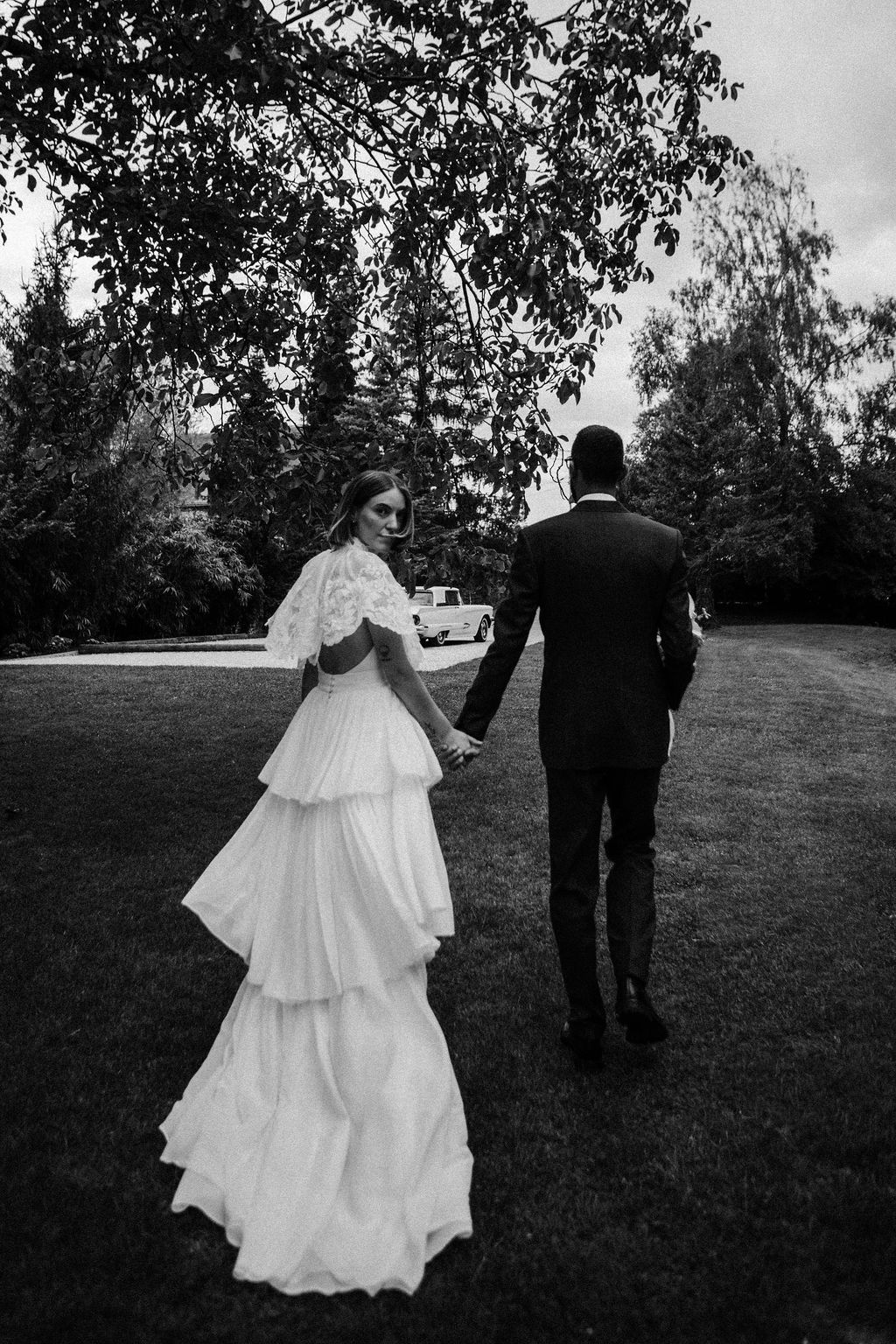 Black and white photo of bride in tiered gown and groom walking hand in hand across a garden lawn.