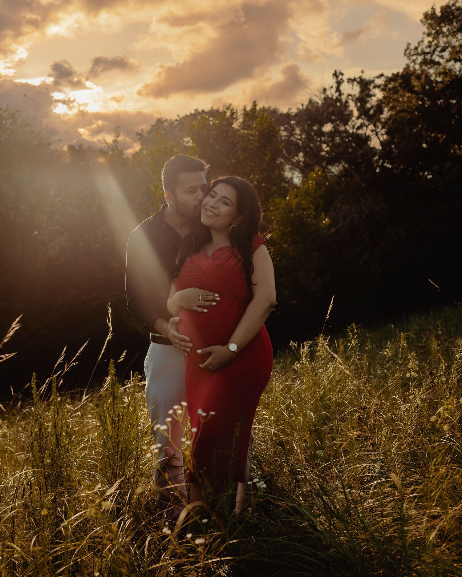 Silhouetted couple embraces in golden sunlit field during sunset while wind blows through tall grass.
