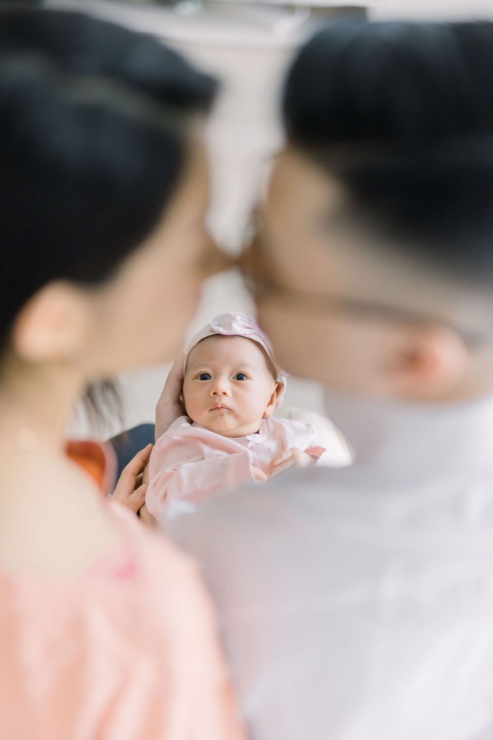 A baby wearing a pink outfit and white hat looks up with curiosity while being held by an adult in a blurred background.