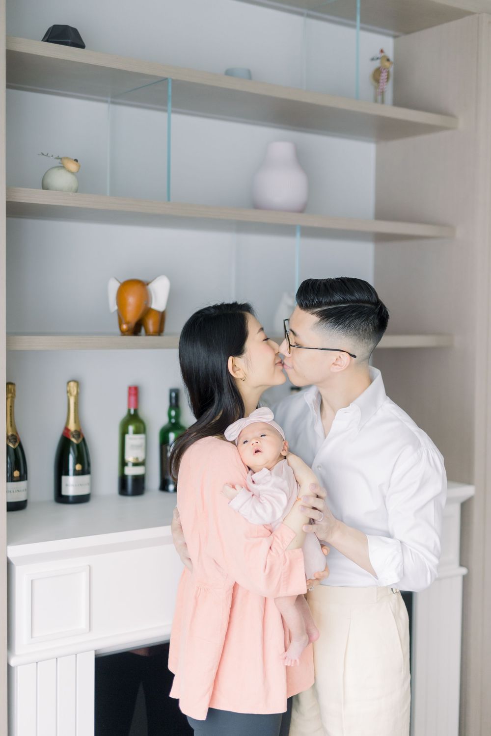 A couple embraces in a modern kitchen with white shelves displaying wine bottles and decorative items in the background.