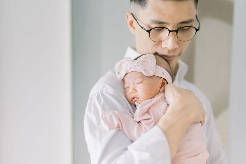 Parent and baby in white outfits share tender moment while sitting together on white couch.