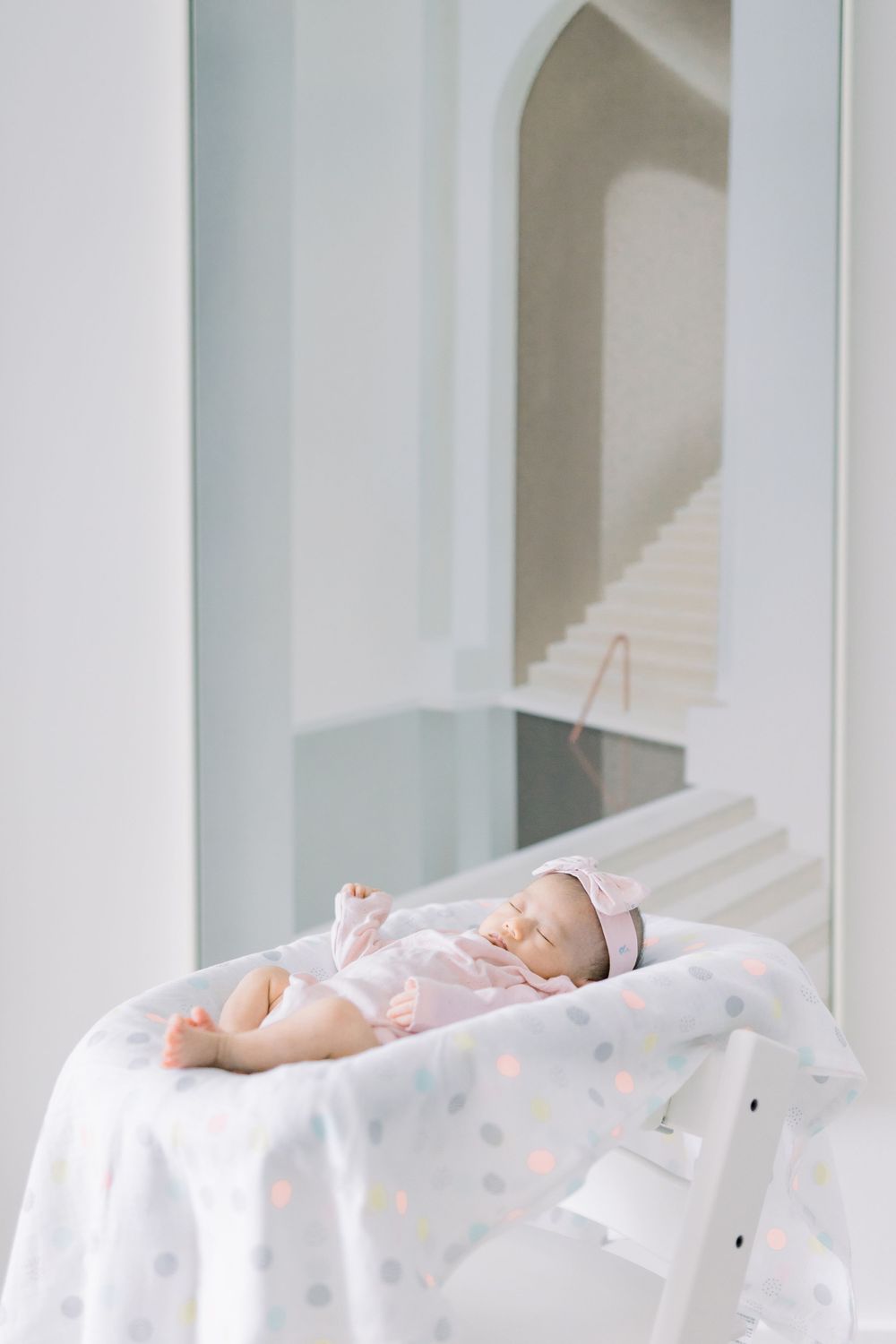 A sleeping baby in white polka dot pajamas lies peacefully on white bedding with natural light streaming through windows.