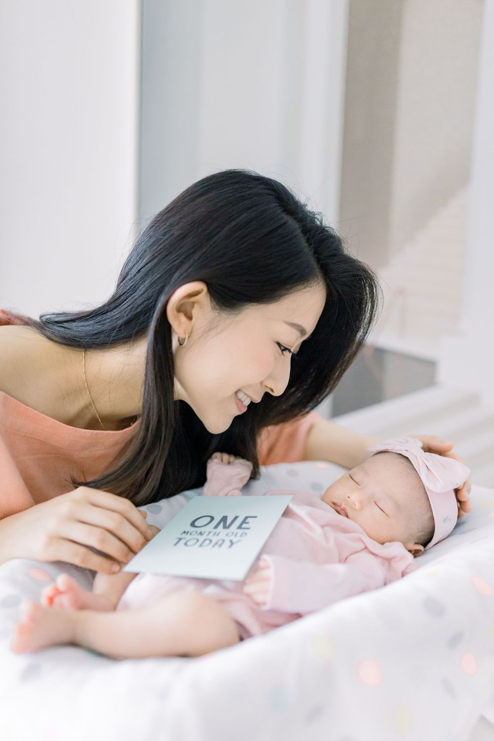 A tender moment between mother and newborn baby lying on white bedding in a bright room.