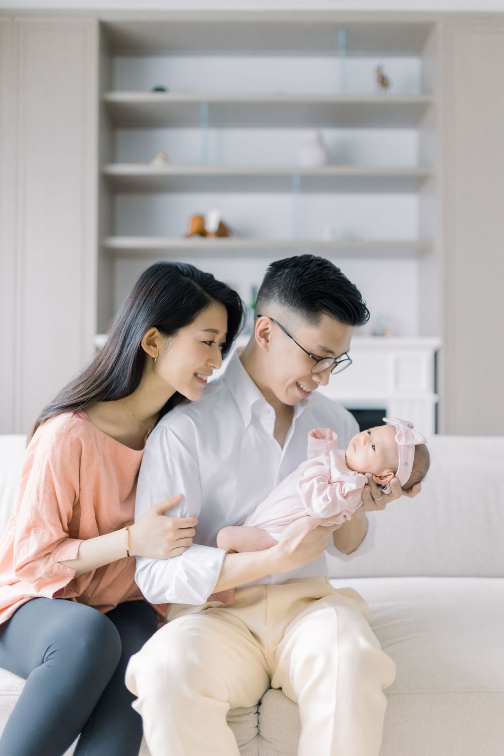 Parents sitting on white couch in living room with newborn baby wearing pink outfit.
