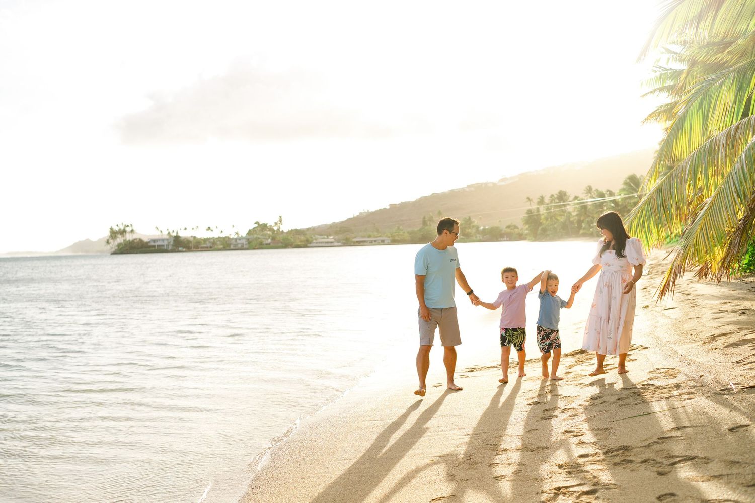 A family walks together holding hands along a tropical beach at sunset as waves lap at the shore. Oahu family photographer