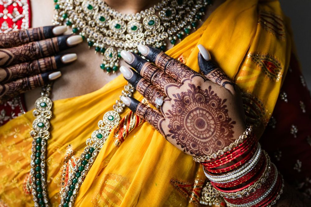 Detailed mehndi art on hands adorned with bangles and traditional Indian bridal jewelry against yellow fabric.