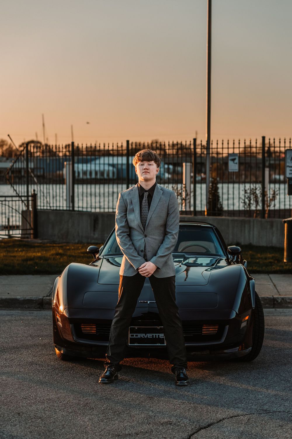 Young man in gray blazer posing against a black Corvette at sunset in a parking lot.