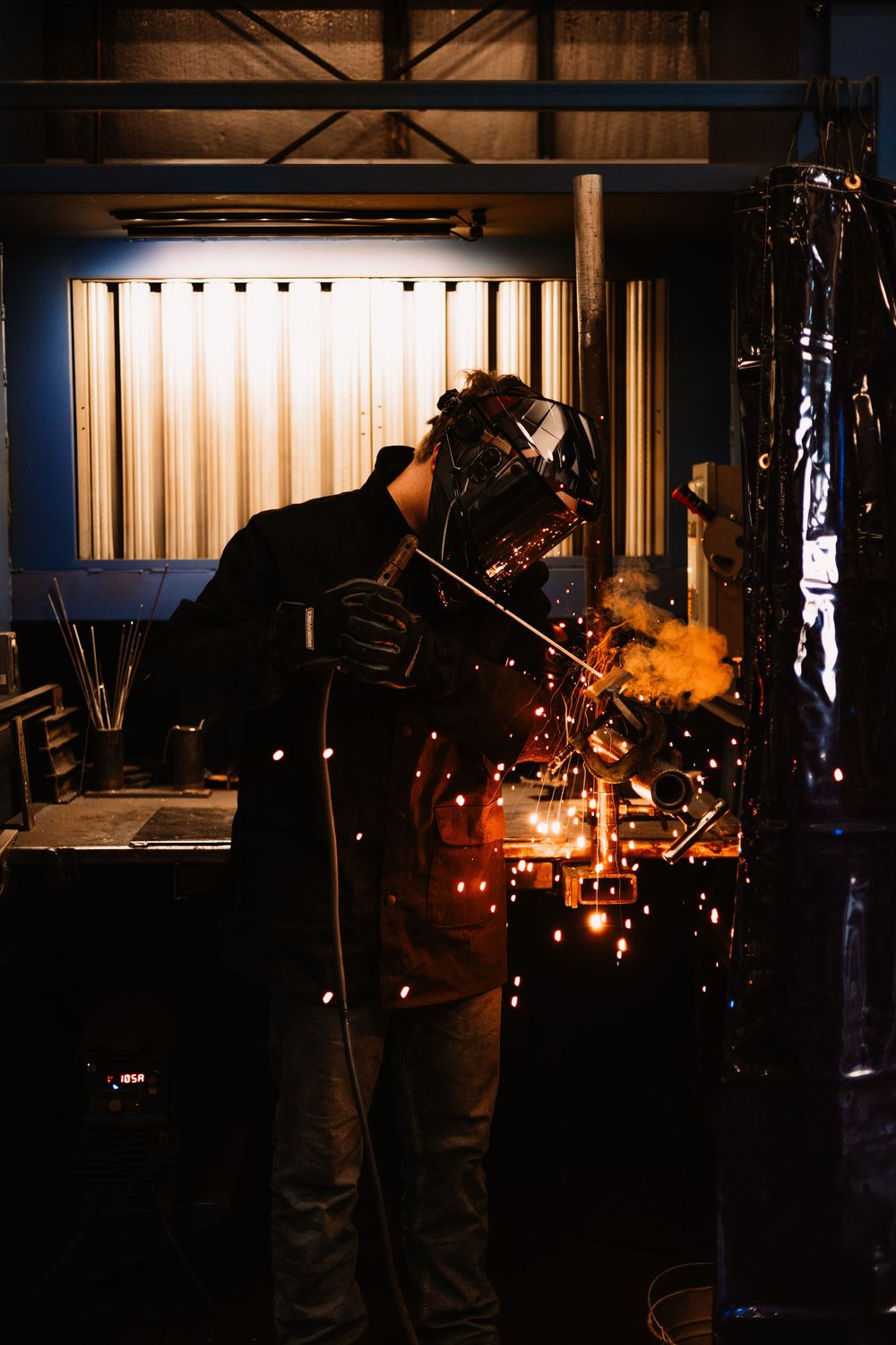 A welder in protective gear creates bright sparks while working with metal in a dark industrial workshop.