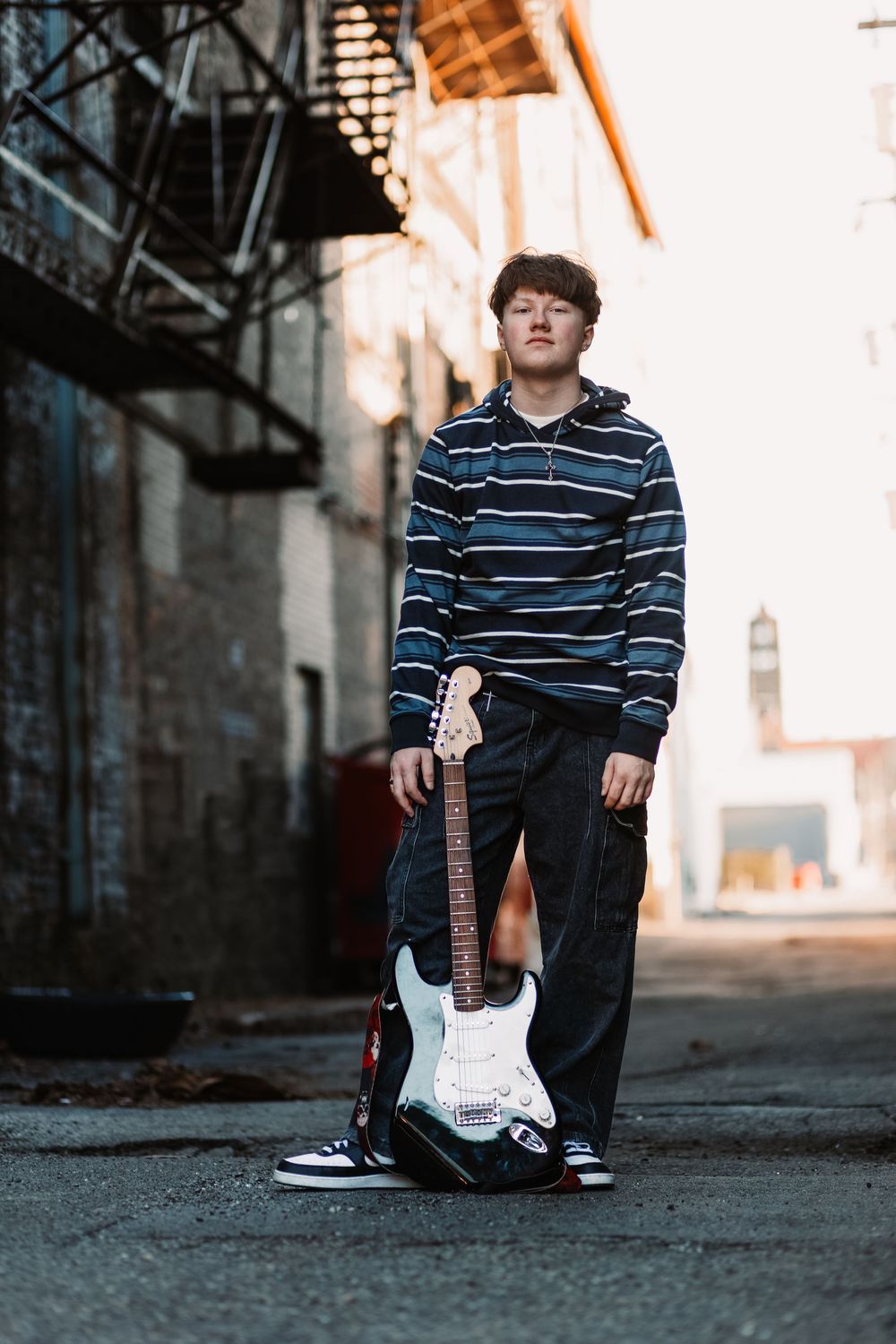 Young musician stands in urban alley holding electric guitar, wearing striped hoodie and black jeans.