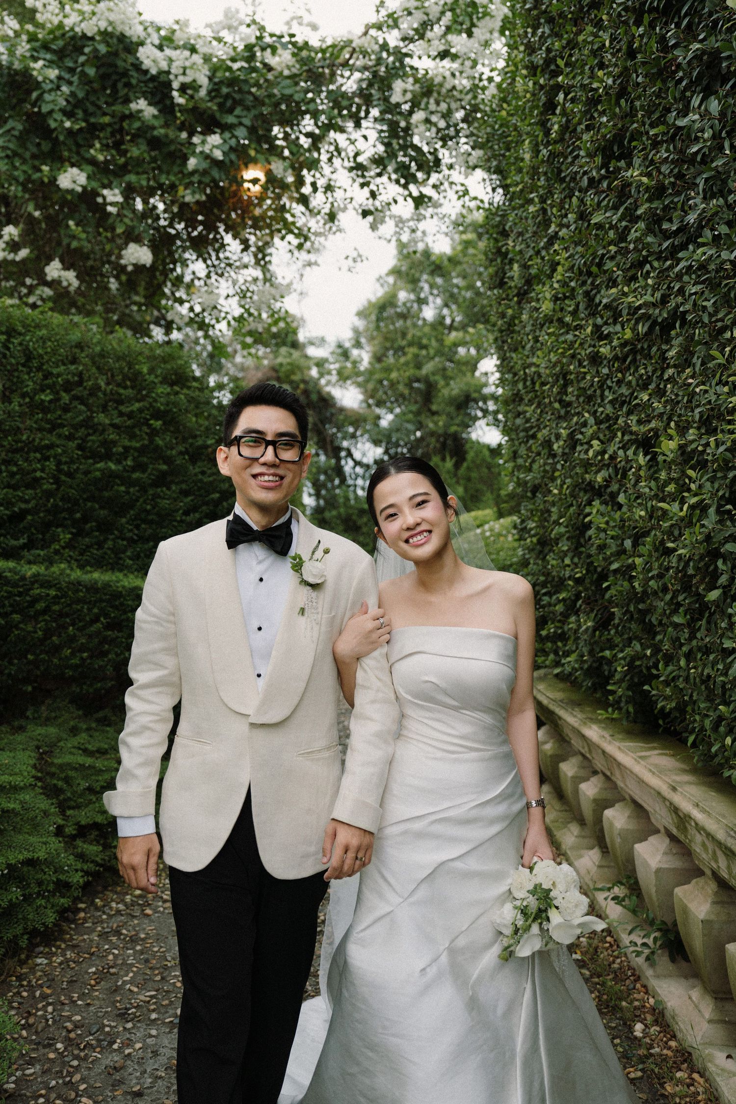Wedding couple poses in formal attire against lush garden backdrop with natural lighting.