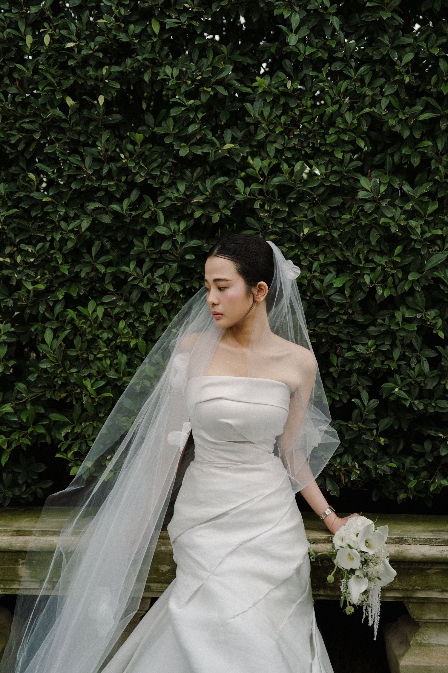 Elegant bride in strapless gown and flowing veil poses in front of manicured garden hedge.