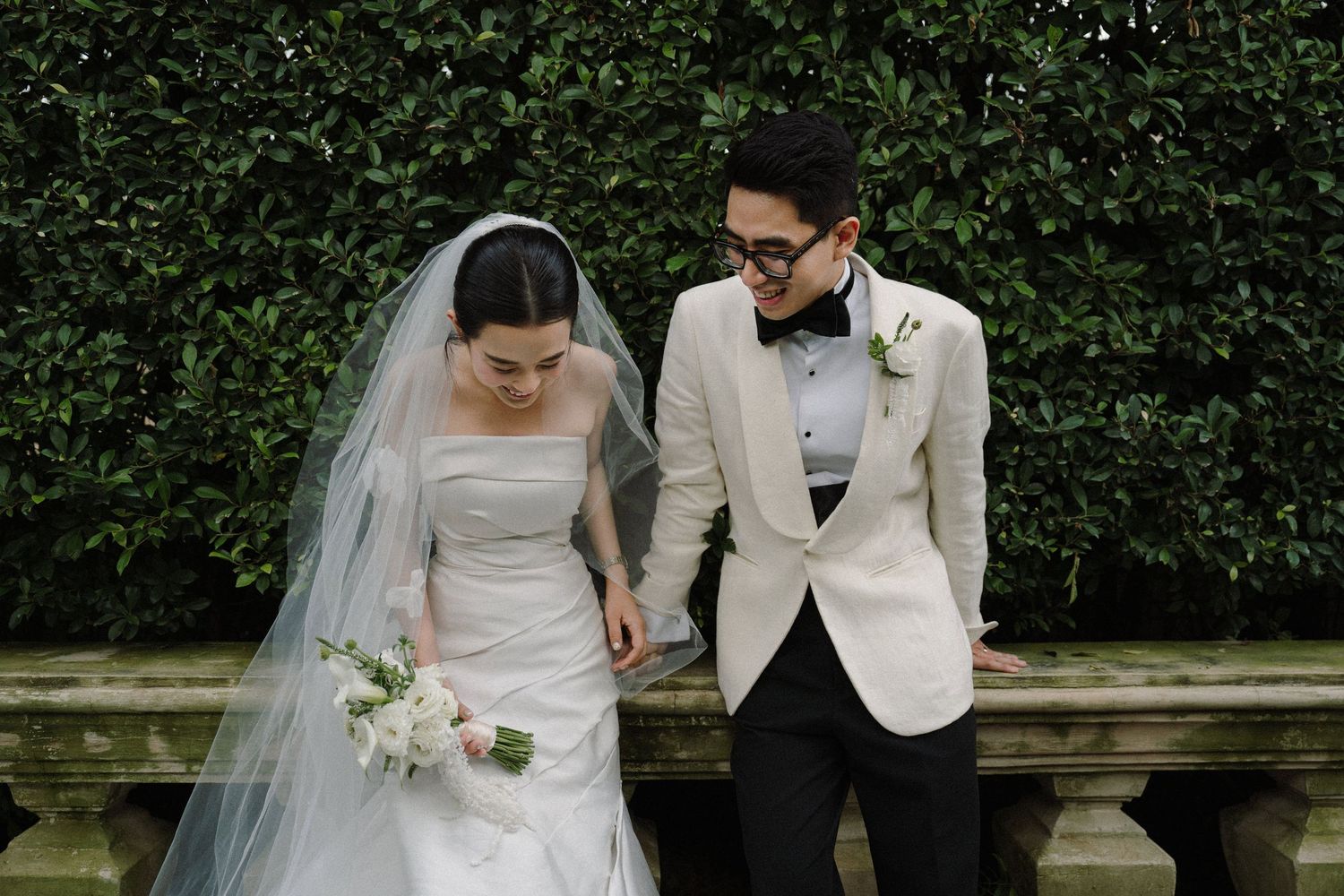 Wedding couple walking and smiling together along a garden path, with the bride in white and groom in cream jacket.