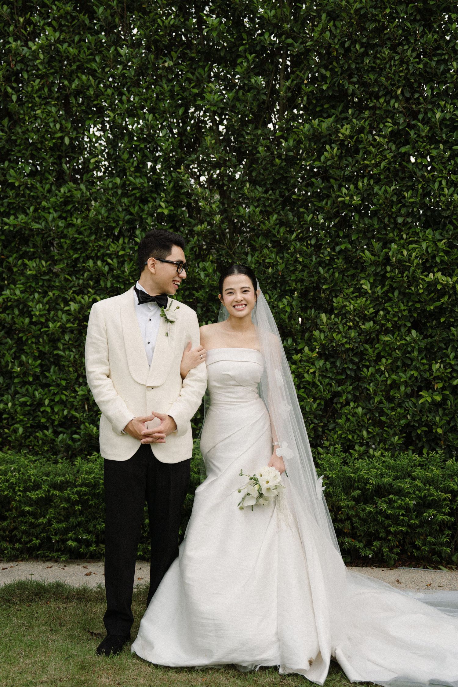 Wedding couple posing together in front of a manicured hedge wall during their outdoor photoshoot.