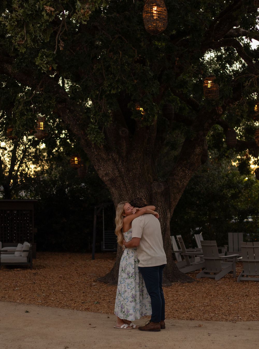A romantic moment between a couple in a garden setting with string lights hanging from trees at dusk.